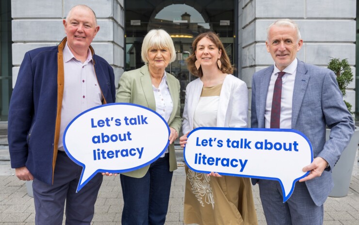 Two men and two women standing on front of building holding signs that say 'Let's talk about literacy' in blue text on white speech bubble.