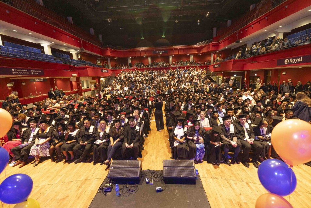 A large group of Mayo College of FET graduates in black caps and gowns sit in rows at their graduation ceremony inside a spacious auditorium. The audience fills the seats behind them, and the venue is decorated with orange and blue balloons, creating a festive atmosphere.