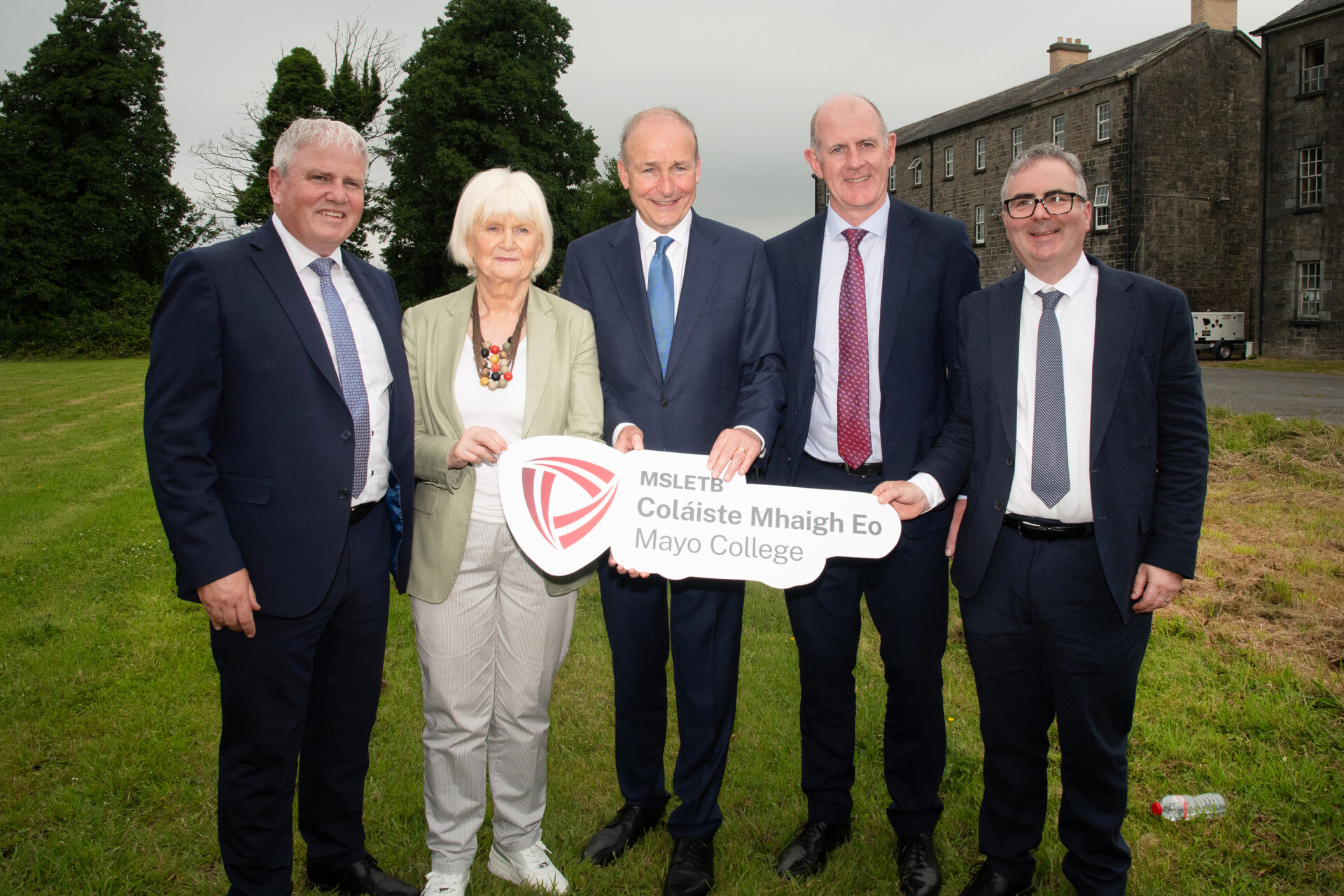Four men in suits and one woman, standing on grass holding sign that says: Mayo College on it.