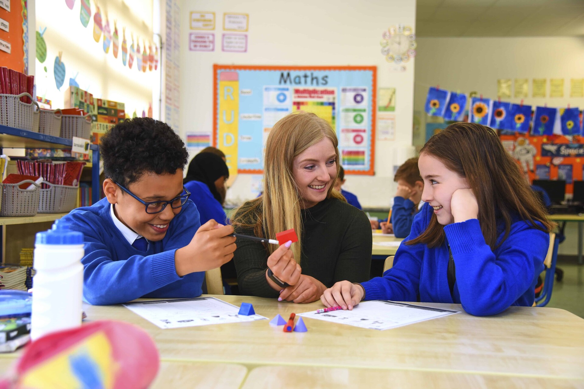 Two students smiling as a teacher helps them during a classroom lesson.