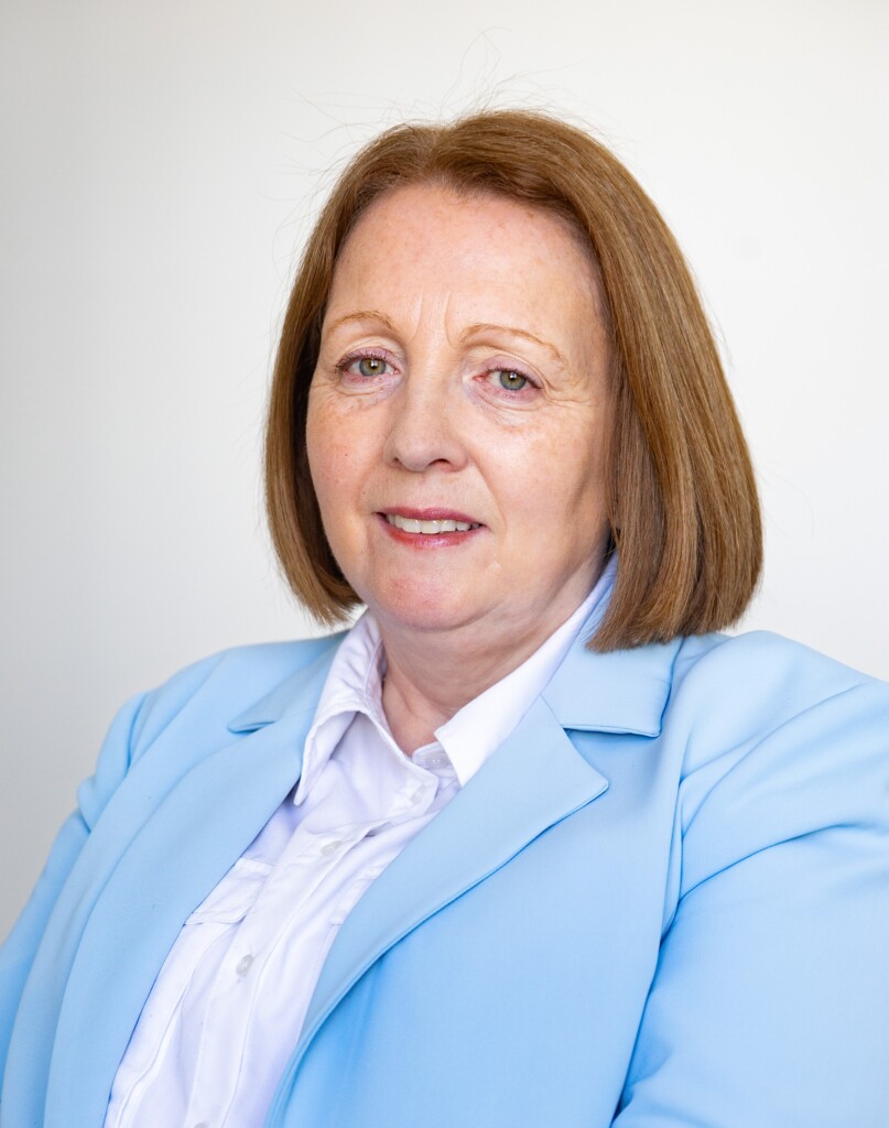 A professional headshot of a woman with shoulder-length auburn hair, wearing a light blue blazer over a white blouse. She is facing the camera with a slight smile against a plain light gray background.