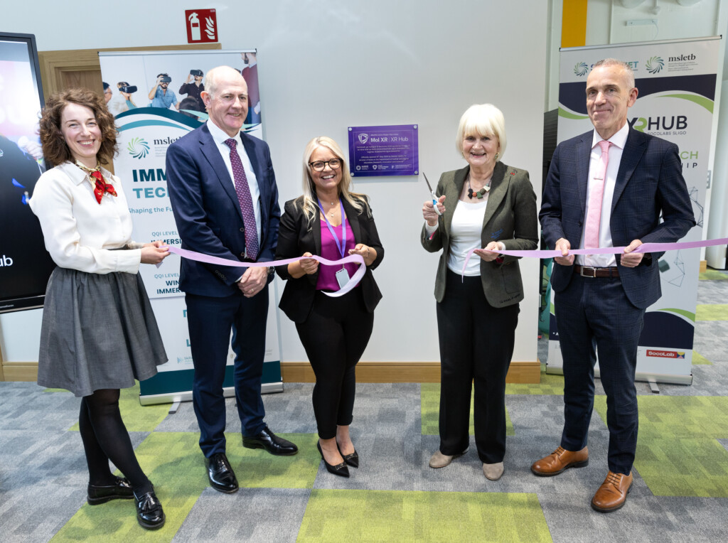 Five people stand together at the official opening of the XR Hub Sligo, holding a pink ribbon during a ribbon-cutting ceremony. One woman is holding scissors, ready to cut the ribbon, while the group smiles for the camera. The event takes place in a modern indoor venue with XR Hub and immersive technology promotional banners displayed in the background, along with a commemorative plaque marking the launch.